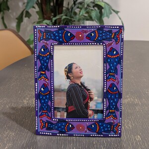 May include: A rectangular picture frame with a purple painted border featuring hand-painted blue fish with red and yellow accents. The frame contains a photograph of a person wearing a black patterned top and gold jewelry, looking upward.