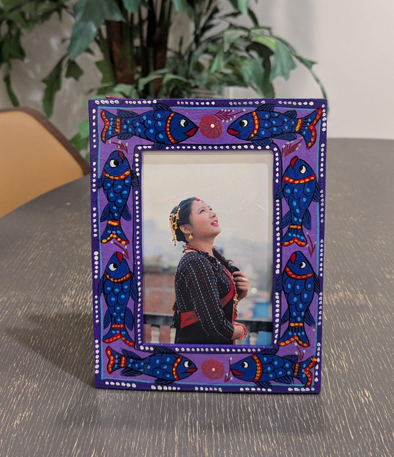 May include: A rectangular picture frame with a purple painted border featuring hand-painted blue fish with red and yellow accents. The frame contains a photograph of a person wearing a black patterned top and gold jewelry, looking upward.