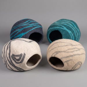 May include: Four felted pet caves in shades of blue, beige, and gray. Each cave has a circular opening and a striped pattern. The caves are designed for small pets and are made of soft, natural materials.