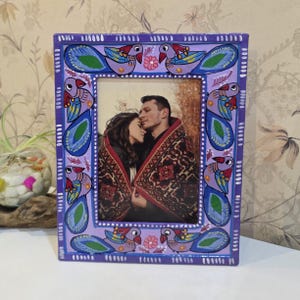 May include: A rectangular picture frame with a light purple base, hand-painted with a repeating pattern of stylised birds in shades of blue, green, and red. The frame features small white dot accents and floral motifs, surrounding a photograph of two people.