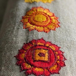 May include: A cylindrical bolster pillow made of light brown, textured linen fabric. The surface features four embroidered circular motifs arranged in a vertical line, showcasing vibrant red, orange, yellow, and green thread colors.