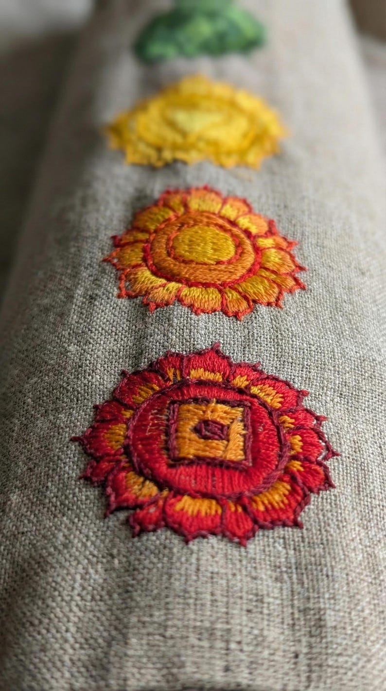 May include: A cylindrical bolster pillow made of light brown, textured linen fabric. The surface features four embroidered circular motifs arranged in a vertical line, showcasing vibrant red, orange, yellow, and green thread colors.