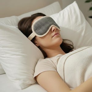 May include: A person wearing a grey and cream sleep mask with a herringbone pattern, resting on a white pillow. The mask is designed to block out light for better sleep. The person is covered with a white blanket.