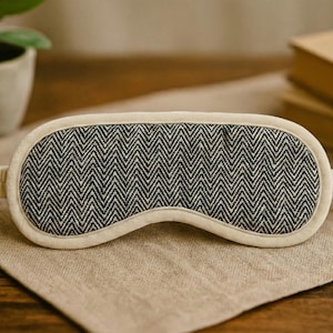 May include: A sleep mask featuring a black and white herringbone design, with a cream-coloured trim and straps. It is placed on a beige linen surface. A small potted plant and books are in the background.