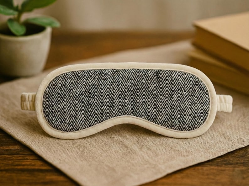 May include: A sleep mask featuring a black and white herringbone design, with a cream-coloured trim and straps. It is placed on a beige linen surface. A small potted plant and books are in the background.
