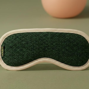 May include: A sleep mask with a dark green center and a cream-colored border and straps. The mask has a textured appearance and is designed to block out light. The background is a muted green with a peach-colored sphere.