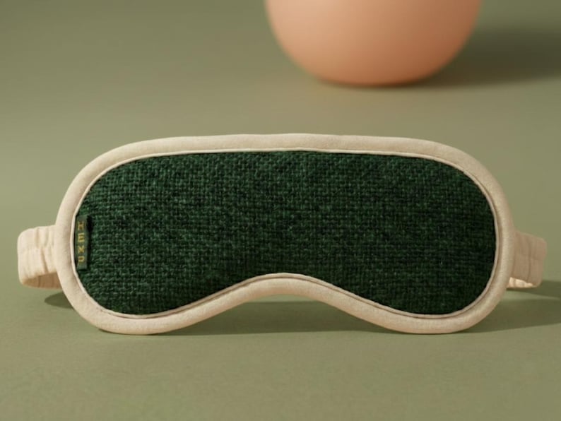 May include: A sleep mask with a dark green center and a cream-colored border and straps. The mask has a textured appearance and is designed to block out light. The background is a muted green with a peach-colored sphere.