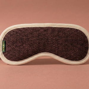 May include: A brown sleep mask with a textured fabric center and a cream-colored border and strap. The mask has a curved shape to fit the eyes and is designed to block out light. A small green tag is attached to the mask.