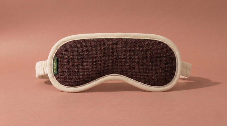 May include: A brown sleep mask with a textured fabric center and a cream-colored border and strap. The mask has a curved shape to fit the eyes and is designed to block out light. A small green tag is attached to the mask.