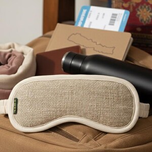 May include: A tan backpack filled with travel essentials. Items include a rolled blanket, a sleep mask, a black water bottle, a passport, and a boarding pass. The sleep mask is beige with a woven texture.