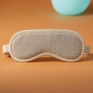 May include: A beige sleep mask with a herringbone pattern. The mask has a soft, padded design with a matching fabric-covered band. The mask is designed to block out light for better sleep. The background is a solid orange colour.