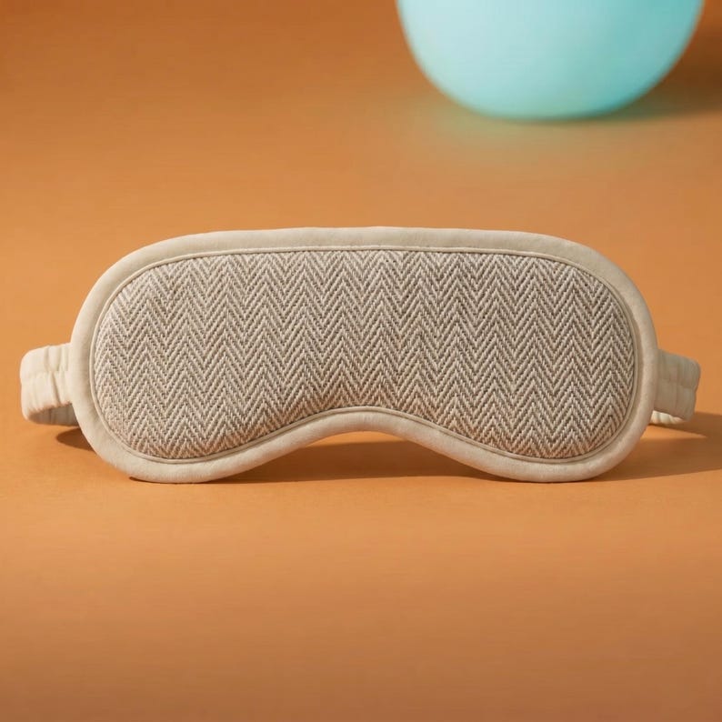 May include: A beige sleep mask with a herringbone pattern. The mask has a soft, padded design with a matching fabric-covered band. The mask is designed to block out light for better sleep. The background is a solid orange colour.