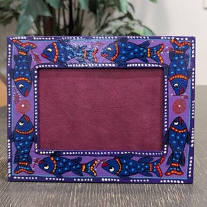 May include: A rectangular purple picture frame featuring a hand-painted design of blue fish with orange and yellow accents. The frame has a dark maroon interior border and a series of small white dots along the outer and inner edges of the frame.