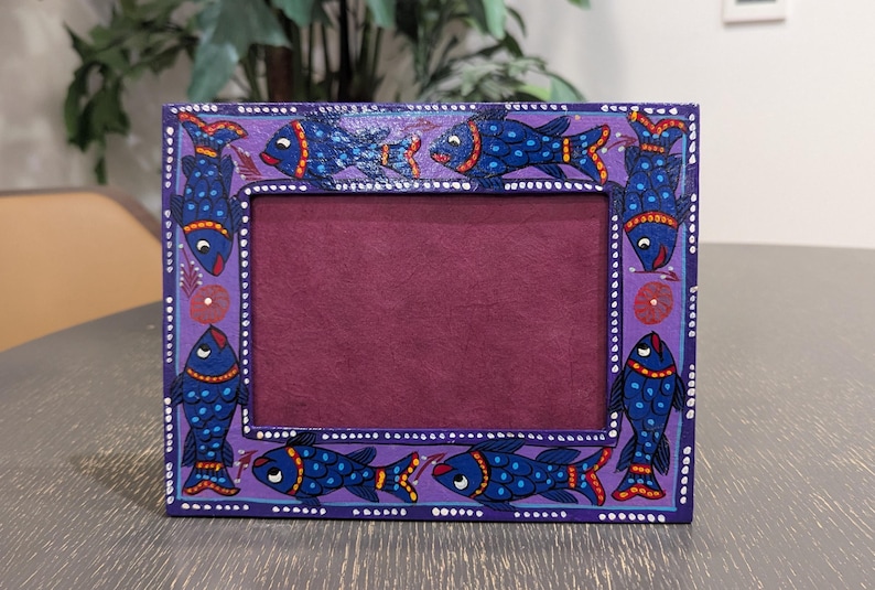 May include: A rectangular purple picture frame featuring a hand-painted design of blue fish with orange and yellow accents. The frame has a dark maroon interior border and a series of small white dots along the outer and inner edges of the frame.