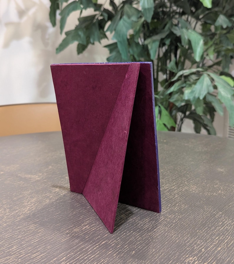 May include: A rectangular picture frame stand featuring a deep burgundy velvet-textured backing. The frame is positioned upright, showcasing the triangular support structure and the dark purple edge of the frame border visible from this side angle.