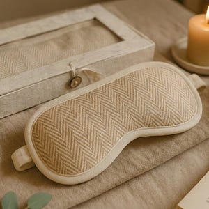 May include: A beige sleep mask with a herringbone pattern and a matching box. The mask is on a neutral-colored surface with a lit candle, a cup, and a book. The mask has a soft, padded design.