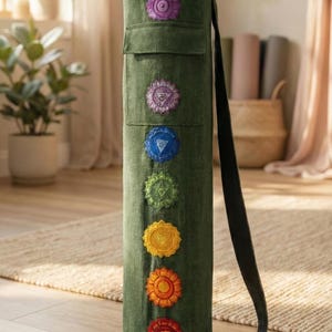 May include: A cylindrical dark green yoga mat bag featuring a vertical row of seven embroidered chakra symbols in purple, blue, green, yellow, orange, and red. The bag includes a long black shoulder strap and a top drawstring closure mechanism.