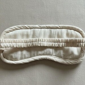May include: A cream-coloured sleep mask with a soft, rounded shape. The mask has a wide, elasticated band for a comfortable fit. The mask is made of a soft, textured fabric, designed to block out light for better sleep.