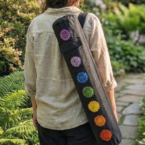 May include: A black cylindrical yoga mat bag featuring seven embroidered chakra symbols in a vertical row, ranging from purple at the top to red at the bottom. The bag has a black shoulder strap and is worn over the shoulder, displaying the colorful circular designs clearly against the dark fabric.