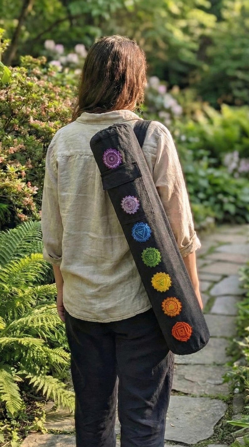 May include: A black cylindrical yoga mat bag featuring seven embroidered chakra symbols in a vertical row, ranging from purple at the top to red at the bottom. The bag has a black shoulder strap and is worn over the shoulder, displaying the colorful circular designs clearly against the dark fabric.