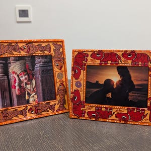 May include: Two rectangular picture frames with hand-painted borders. The left frame features orange borders with brown fish illustrations. The right frame features orange borders with red elephant illustrations. Both frames hold photographs.