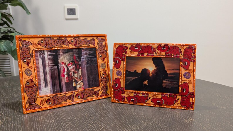 May include: Two rectangular picture frames with hand-painted borders. The left frame features orange borders with brown fish illustrations. The right frame features orange borders with red elephant illustrations. Both frames hold photographs.