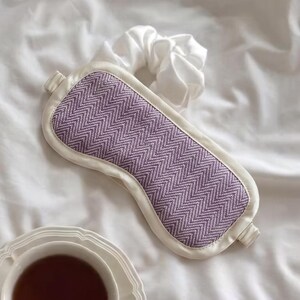 May include: A lavender and white sleep mask with a chevron pattern. The mask is surrounded by a white border and has a white satin scrunchie next to it. A cup of tea sits in the bottom left corner.