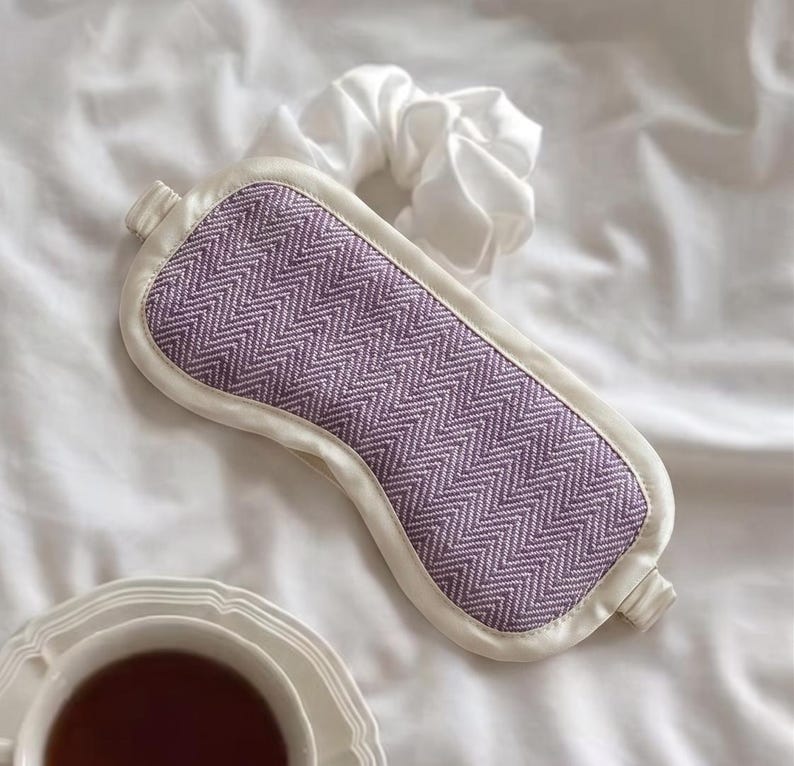 May include: A lavender and white sleep mask with a chevron pattern. The mask is surrounded by a white border and has a white satin scrunchie next to it. A cup of tea sits in the bottom left corner.