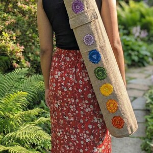 May include: A cylindrical beige yoga mat bag featuring seven embroidered chakra symbols in their traditional colors arranged vertically along the length of the bag, including purple, indigo, blue, green, yellow, orange, and red circular designs.