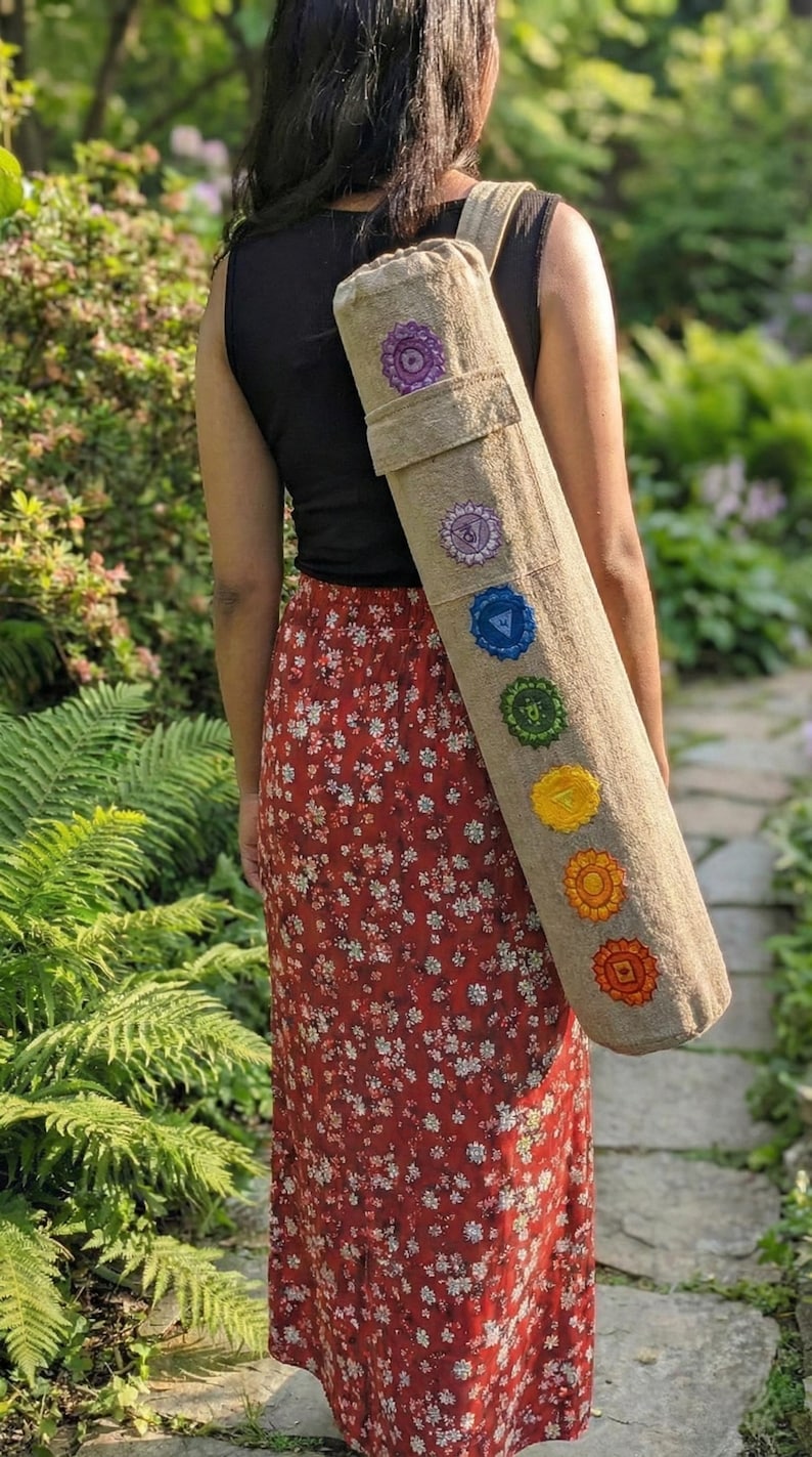 May include: A cylindrical beige yoga mat bag featuring seven embroidered chakra symbols in their traditional colors arranged vertically along the length of the bag, including purple, indigo, blue, green, yellow, orange, and red circular designs.