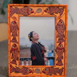 May include: A rectangular orange picture frame featuring hand-painted brown fish motifs along the border. The frame displays a photograph of a person looking upward, set against an outdoor background. The border includes small white dot accents.