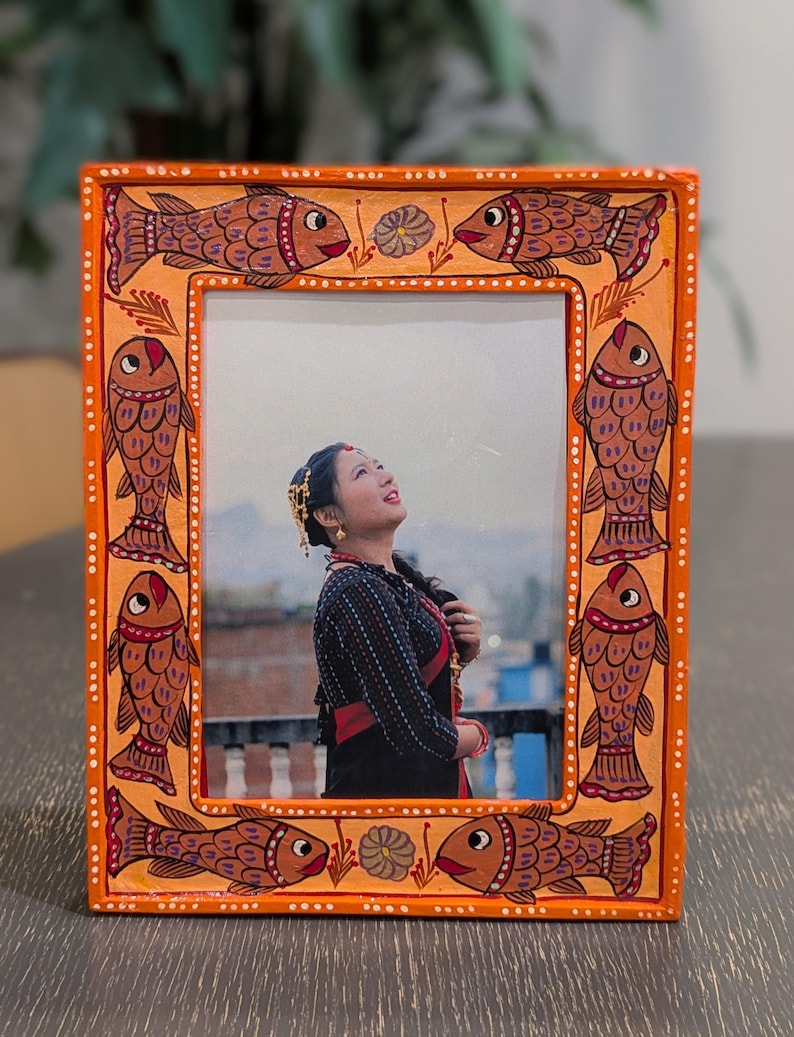May include: A rectangular orange picture frame featuring hand-painted brown fish motifs along the border. The frame displays a photograph of a person looking upward, set against an outdoor background. The border includes small white dot accents.