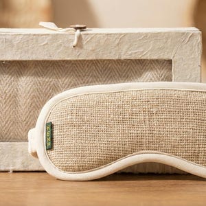 May include: A beige sleep mask with a woven texture, resting on a wooden surface next to a matching box. The mask has a curved shape and a soft, off-white trim. The box has a window revealing a herringbone pattern.