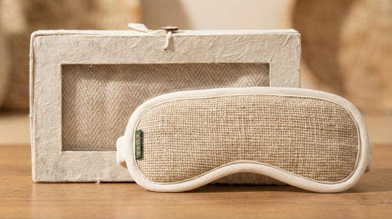 May include: A beige sleep mask with a woven texture, resting on a wooden surface next to a matching box. The mask has a curved shape and a soft, off-white trim. The box has a window revealing a herringbone pattern.