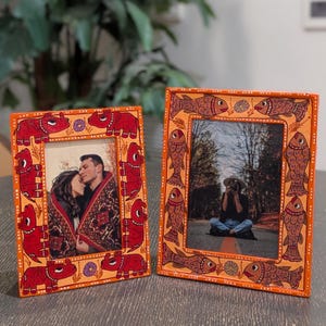 May include: Two rectangular picture frames with orange borders. One frame features red hippopotamus designs, the other has fish. The frames hold photographs of people. The frames are on a gray surface.