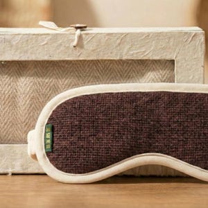 May include: A brown sleep mask with a cream-colored border and a matching box. The mask is made of a textured fabric and has a small green tag. The box is made of a light-colored, textured material.