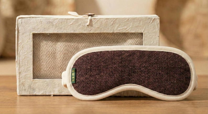 May include: A brown sleep mask with a cream-colored border and a matching box. The mask is made of a textured fabric and has a small green tag. The box is made of a light-colored, textured material.