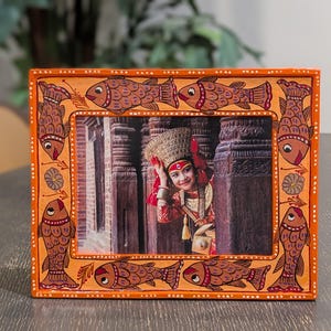 May include: A rectangular orange picture frame featuring hand-painted fish motifs along the border. The frame contains a photograph of a person wearing a traditional headpiece and red clothing, positioned between two dark stone pillars in a temple.