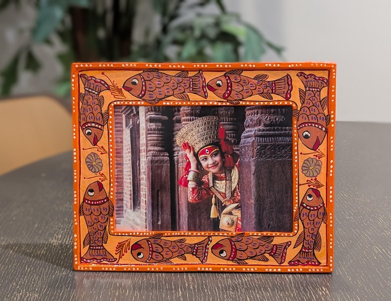 May include: A rectangular orange picture frame featuring hand-painted fish motifs along the border. The frame contains a photograph of a person wearing a traditional headpiece and red clothing, positioned between two dark stone pillars in a temple.