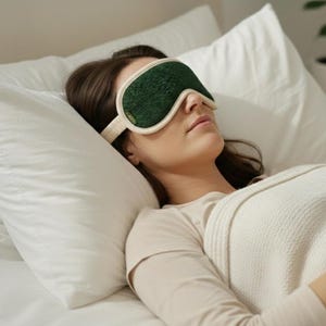 May include: A person wearing a green and cream-colored sleep mask, resting on a white pillow. The mask has a textured green surface and a cream-colored band. The person is covered with a white blanket.