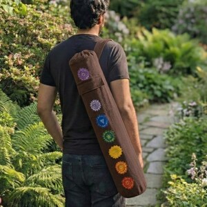 May include: A brown cylindrical yoga mat bag with a shoulder strap, featuring seven embroidered circular chakra symbols in purple, indigo, blue, green, yellow, orange, and red arranged vertically along the length of the fabric carrier.
