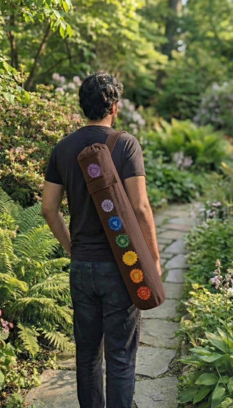 May include: A brown cylindrical yoga mat bag with a shoulder strap, featuring seven embroidered circular chakra symbols in purple, indigo, blue, green, yellow, orange, and red arranged vertically along the length of the fabric carrier.