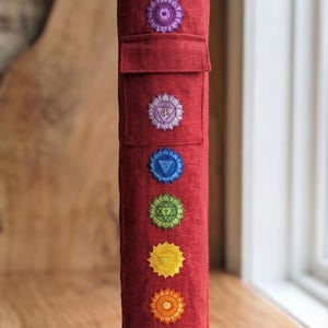 May include: A cylindrical red bolster pillow featuring seven embroidered chakra symbols arranged vertically in a line. The fabric has a visible linen-like texture, and the top section includes a rectangular flap pocket detail above the symbols.
