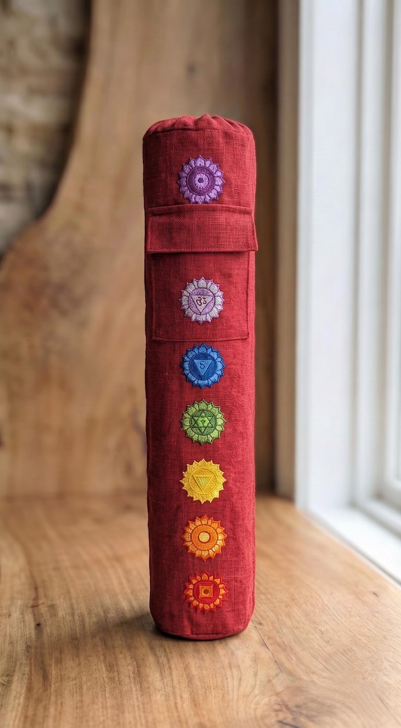 May include: A cylindrical red bolster pillow featuring seven embroidered chakra symbols arranged vertically in a line. The fabric has a visible linen-like texture, and the top section includes a rectangular flap pocket detail above the symbols.