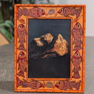 May include: A rectangular orange picture frame featuring hand-painted fish motifs along the border. The frame displays a photograph of a sunlit mountain peak against a dark sky. The border is decorated with white dots and small floral accents.