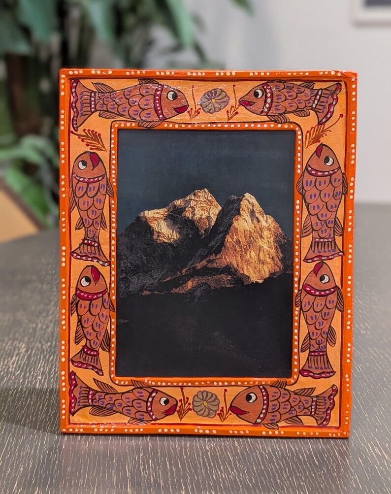 May include: A rectangular orange picture frame featuring hand-painted fish motifs along the border. The frame displays a photograph of a sunlit mountain peak against a dark sky. The border is decorated with white dots and small floral accents.