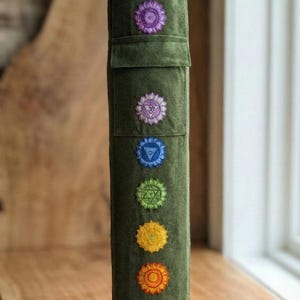 May include: A cylindrical olive green fabric yoga mat bag featuring a vertical row of seven embroidered chakra symbols in purple, light purple, blue, green, yellow, orange, and red. The bag has a drawstring closure at the top and a small pocket.