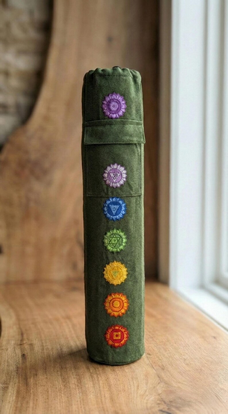 May include: A cylindrical olive green fabric yoga mat bag featuring a vertical row of seven embroidered chakra symbols in purple, light purple, blue, green, yellow, orange, and red. The bag has a drawstring closure at the top and a small pocket.