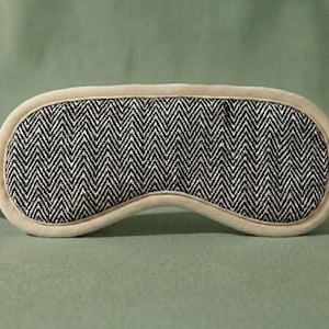 May include: A beige sleep mask featuring a black and white herringbone design. The mask has a soft, padded construction with a matching strap. This eye mask is designed to block out light for improved sleep quality.