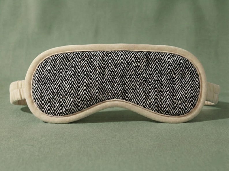 May include: A beige sleep mask featuring a black and white herringbone design. The mask has a soft, padded construction with a matching strap. This eye mask is designed to block out light for improved sleep quality.